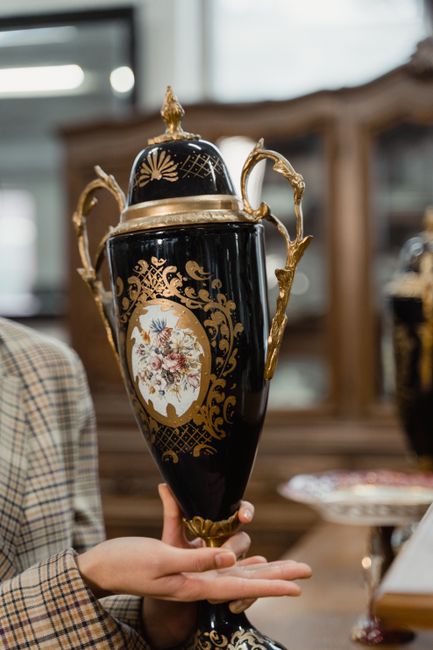 An ornate display of antique silverware on a beige cloth-covered table, including a large decorative bowl with intricate detailing, a polished teapot, and several matching smaller teapots or jugs. Beh